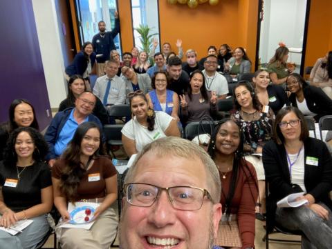 A man with glasses smiling and taking a selfie in front of a group of people in professional attire
