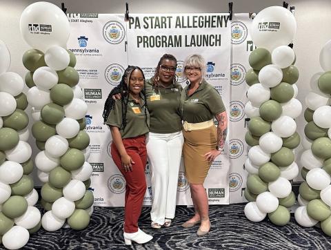 Three women in professional attire pose for a photo in front of a sign that says PA START Allegheny Program Launch with green and white balloons