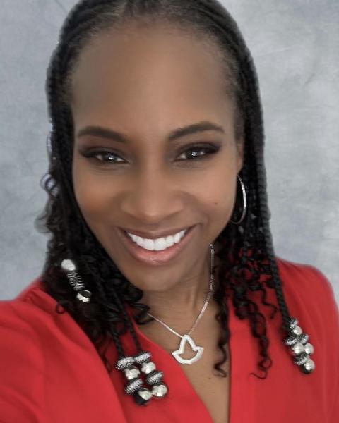 Jenee is a woman with curly black hair put into thin braids with silver beads at the end wearing a red blouse and silver leaf pendant necklace
